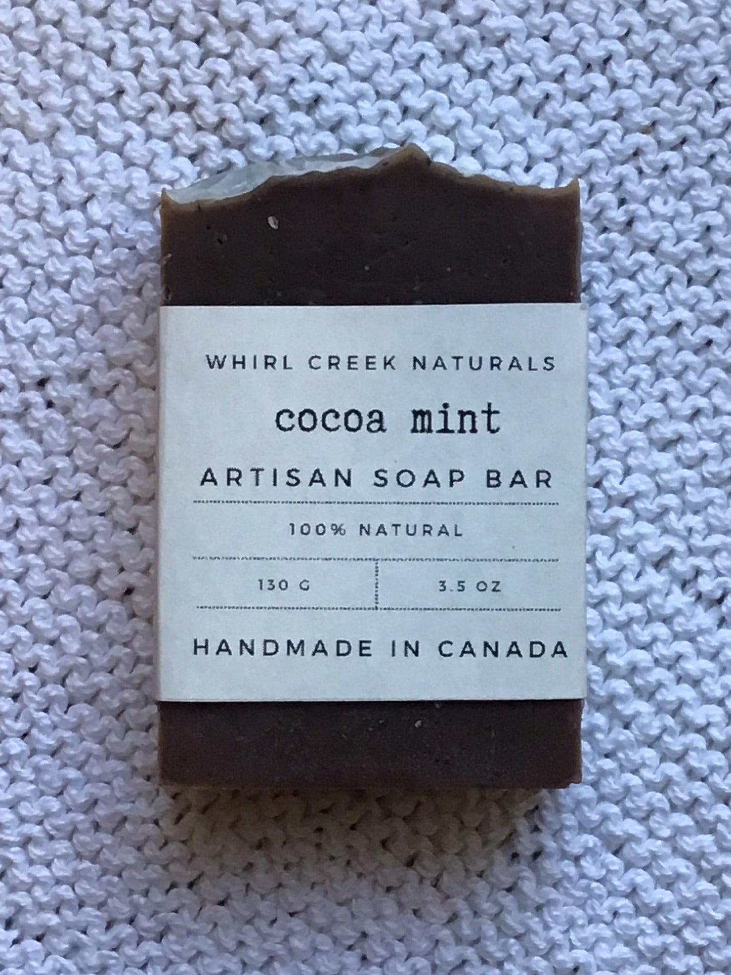 rich dark brown coloured soap bar placed on white cotton washcloth. Paper label wrapped around bar read cocoa mint artisan soap bar 130 grams and hand made in Canada.
