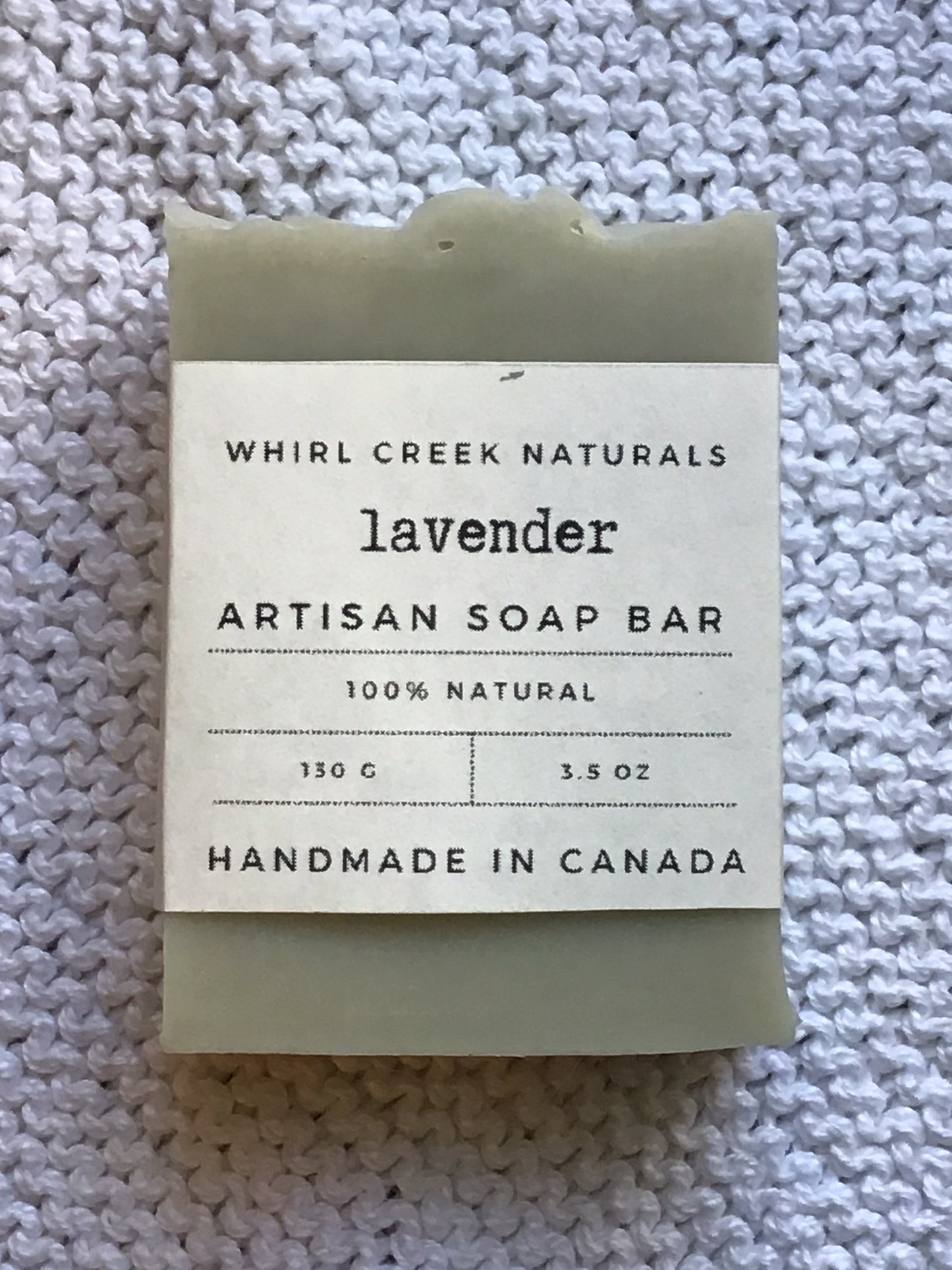 light sage green soap bar placed centered on knit white cotton cloth. Cream-coloured label is wrapped around middle of bar with the words lavender artisan soap bar 130 grams and handmade in Canada written.