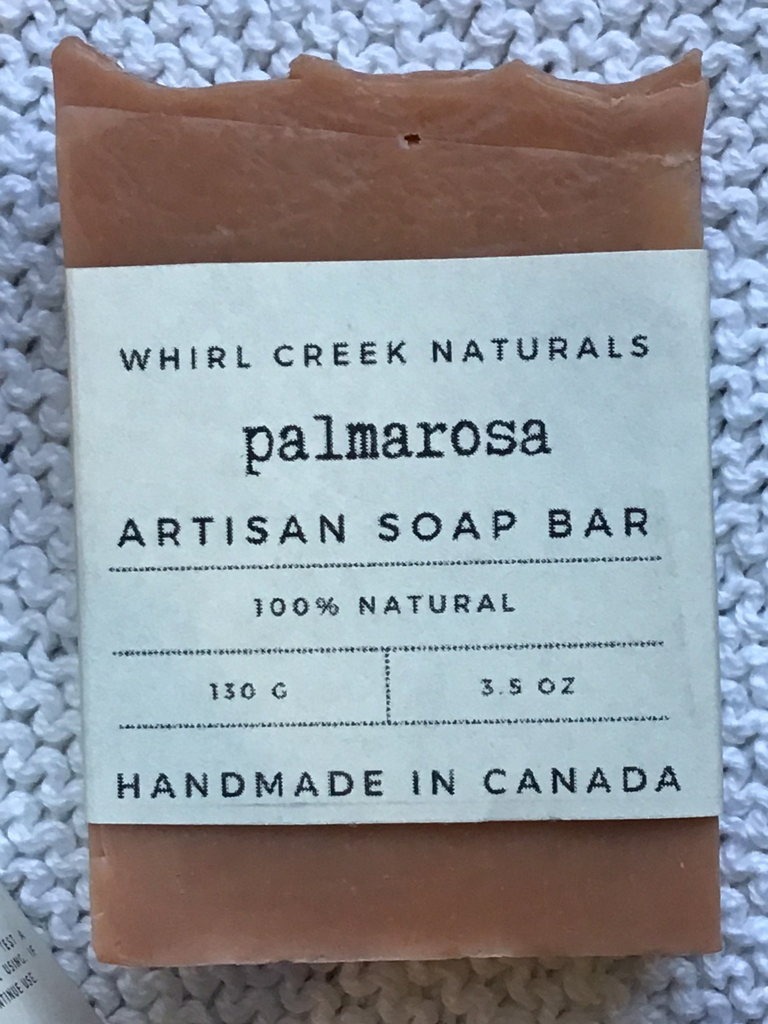 salmon-pink coloured soap bar placed centered on knit white cotton cloth. Cream-coloured label is wrapped around middle of bar with the words palmarosa artisan soap bar 130 grams and handmade in Canada written.