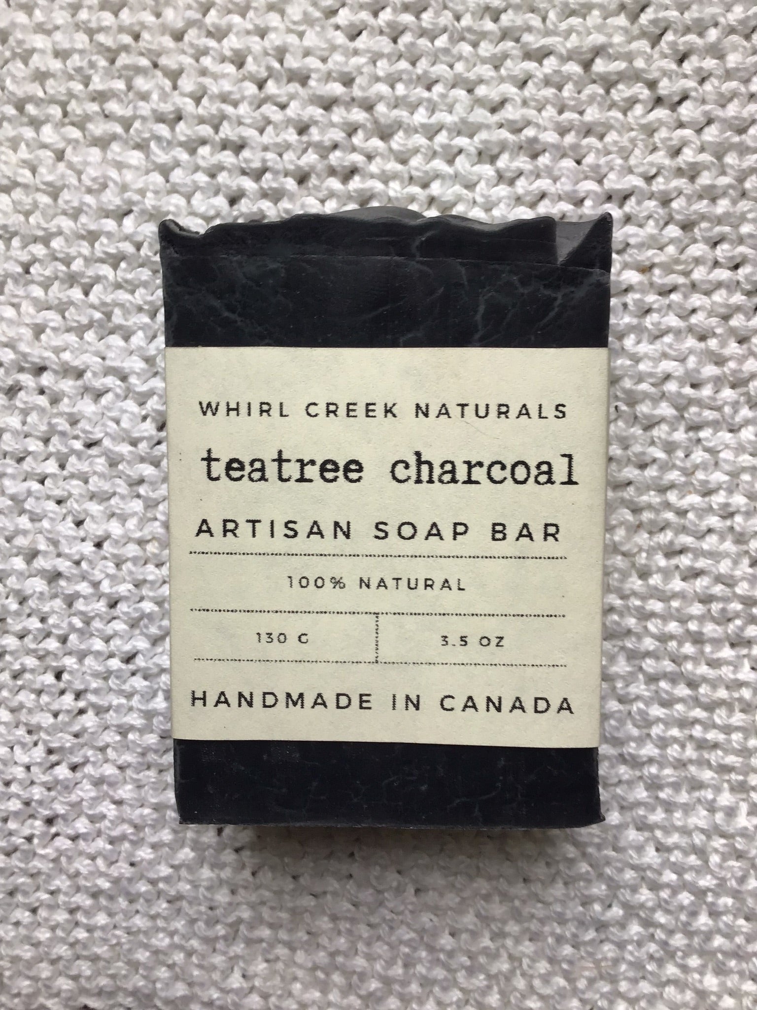 black soap bar placed on white cotton washcloth. Label reads teatree charcoal artisan soap bar 130 grams handmade in Canada