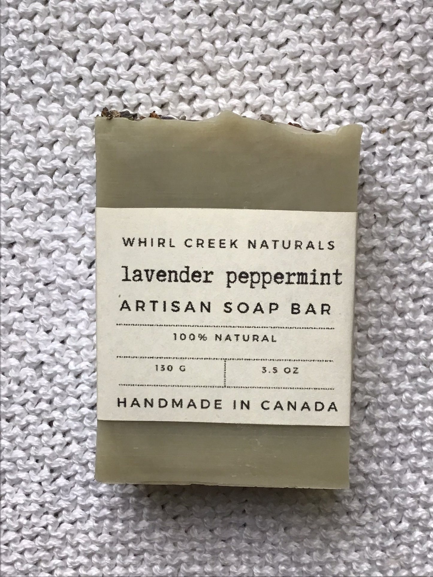 light sage green soap bar placed centered on knit white cotton cloth. Cream-coloured label is wrapped around middle of bar with the words lavender peppermint artisan soap bar 130 grams and handmade in Canada written.