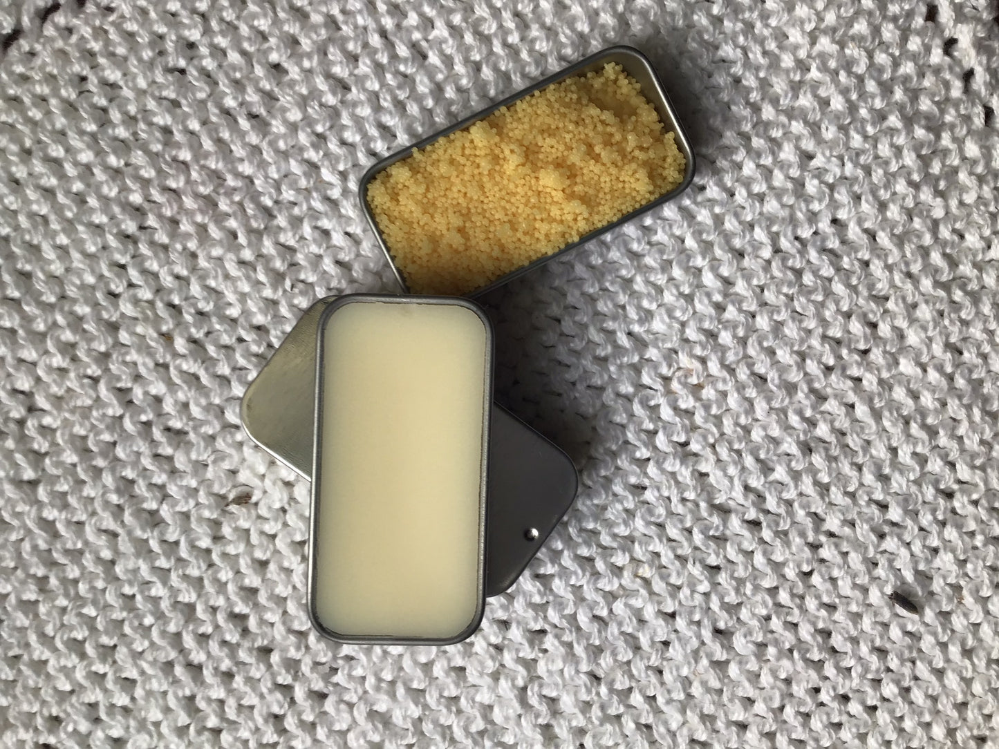 two open rectangular tins placed on knit white cotton cloth. One contains cream-coloured  vegan lip balm, the other a fine yellow granular, Candelilla wax, an plant-based alternative to beeswax. 