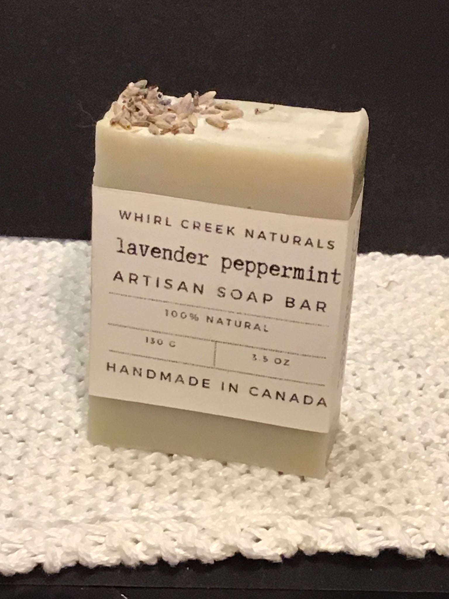 light sage green soap bar placed centered on knit white cotton cloth. Cream-coloured label is wrapped around middle of bar with the words lavender peppermint artisan soap bar 130 grams and handmade in Canada written.