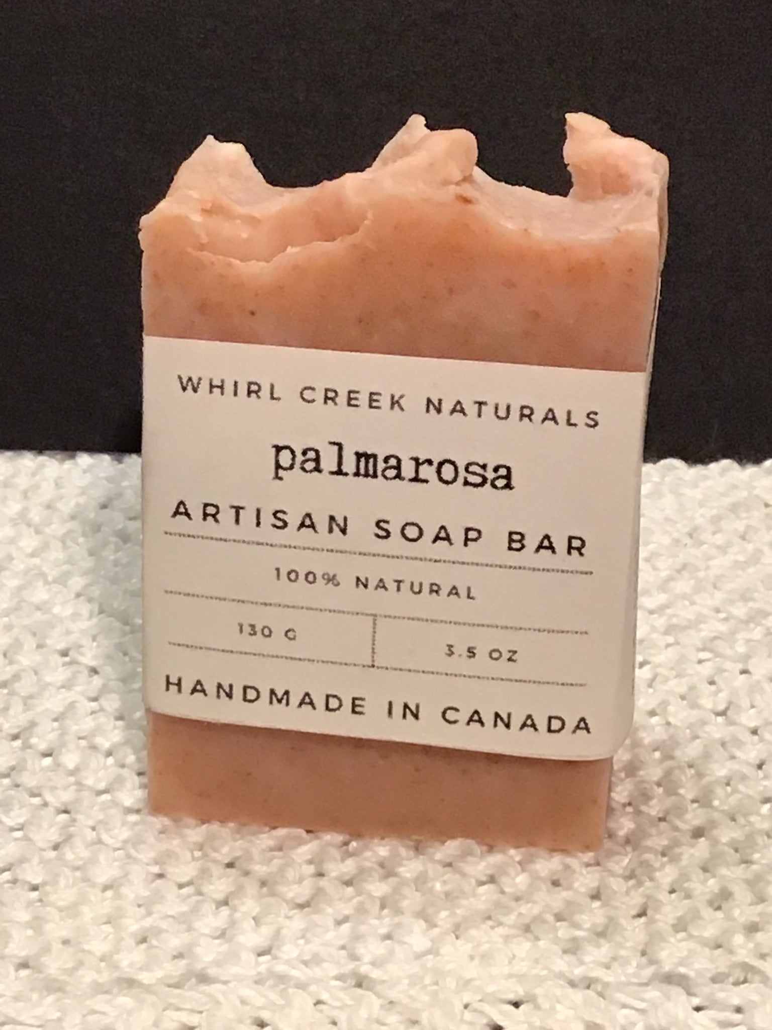 salmon-pink coloured soap bar placed centered on knit white cotton cloth. Cream-coloured label is wrapped around middle of bar with the words palmarosa artisan soap bar 130 grams and handmade in Canada written.