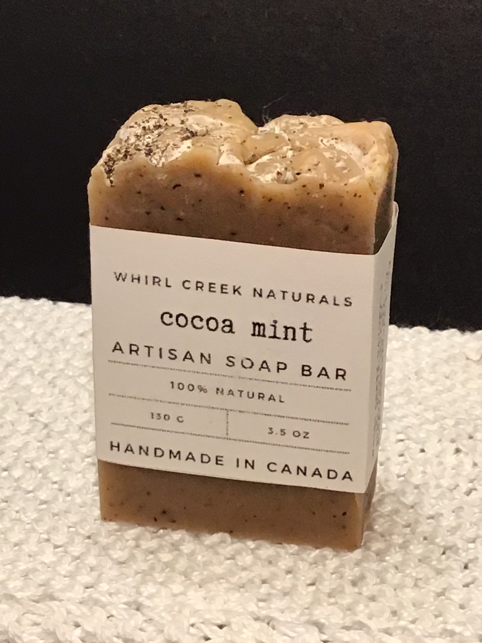 rich dark brown coloured soap bar placed on white cotton washcloth. Paper label wrapped around bar read cocoa mint artisan soap bar 130 grams and hand made in Canada.