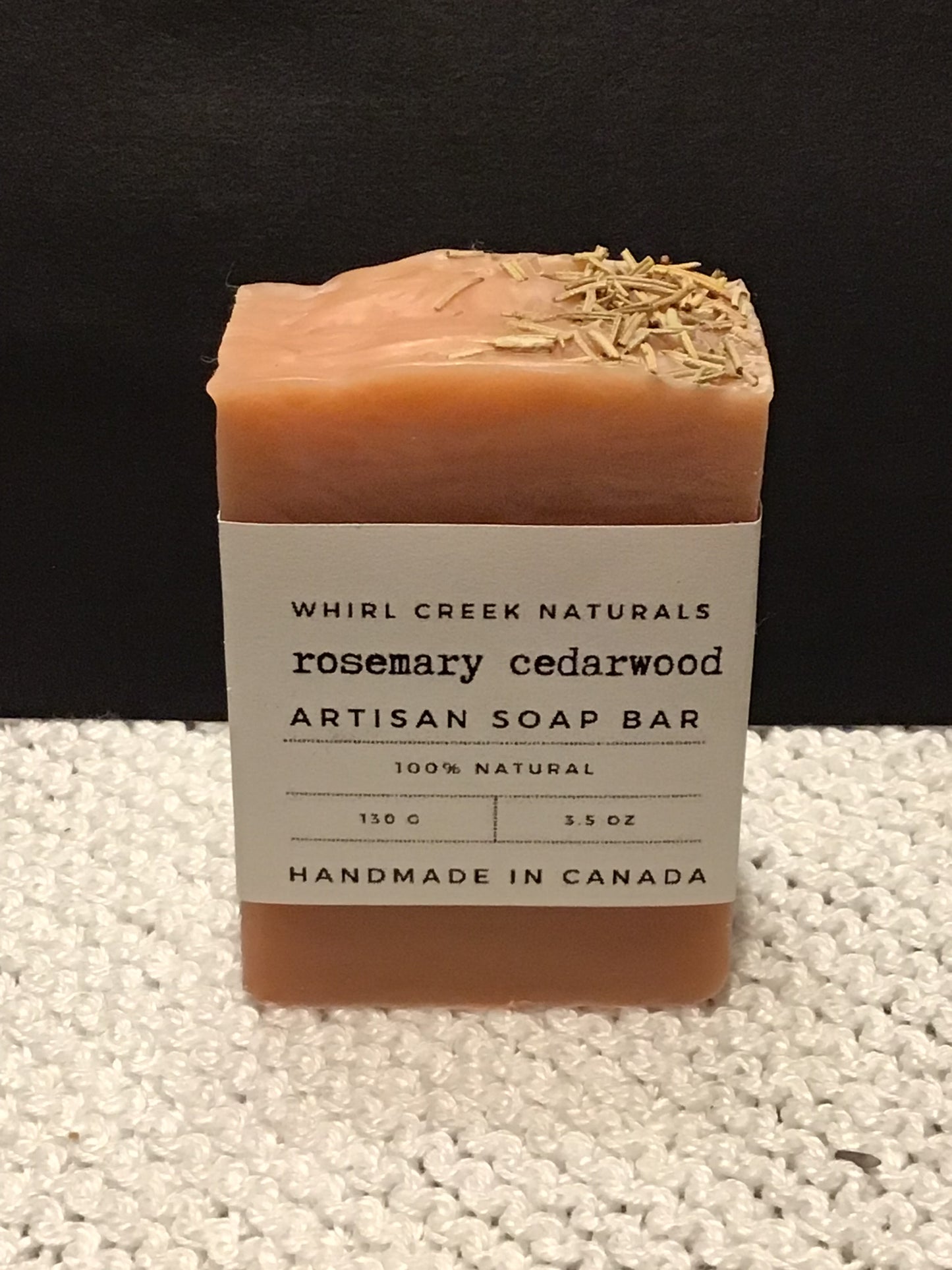 salmon-pink coloured soap bar placed centered on knit white cotton cloth. Cream-coloured label is wrapped around middle of bar with the words cedarwood artisan soap bar 130 grams and handmade in Canada written.