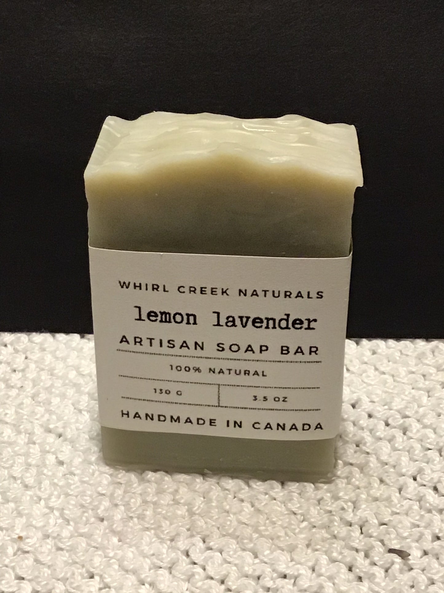 light sage green soap bar placed centered on knit white cotton cloth. Cream-coloured label is wrapped around middle of bar with the words lavender artisan soap bar 130 grams and handmade in Canada written.